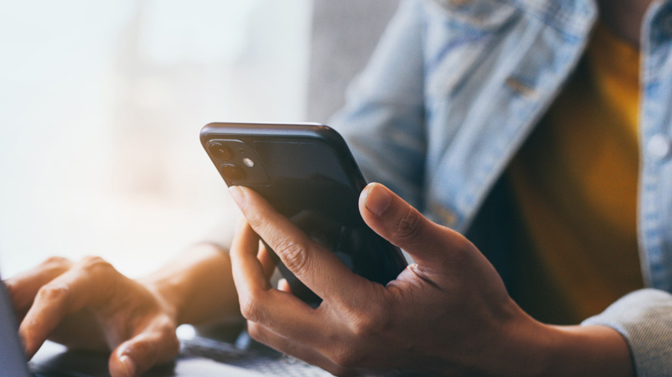 The image shows a close-up of a person s hands holding a smartphone with the screen facing upwards, displaying content on the screen. The background is blurred but appears to be an indoor setting with natural light.