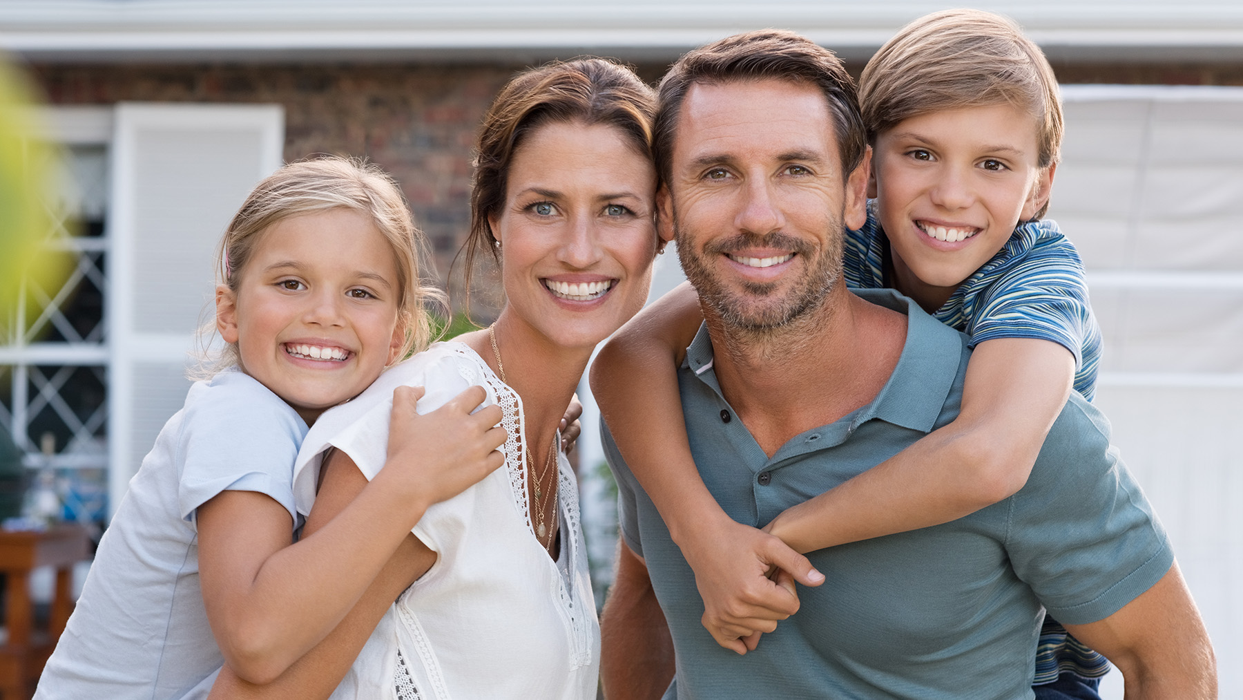 This is a color photograph showing a family of four with two adults and two children posing for a photo outdoors, with a house and fence visible in the background.