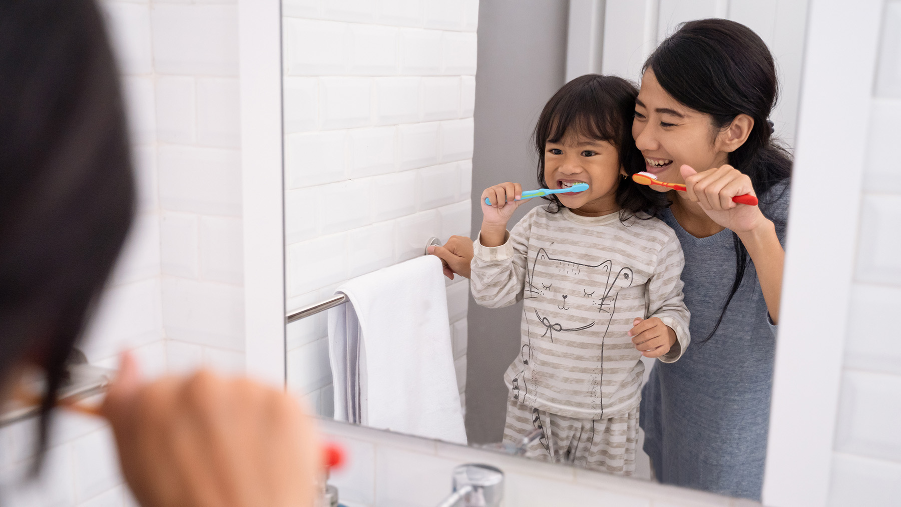 The image shows a woman with a young child in a bathroom setting, both engaged in brushing their teeth at a mirror.