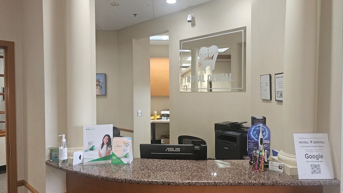 The image shows an interior space, likely a lobby or reception area, with a counter featuring a sign, brochures, and a computer monitor. There are chairs arranged for seating, a large window allowing natural light into the room, and a decorative element on the wall above the counter.