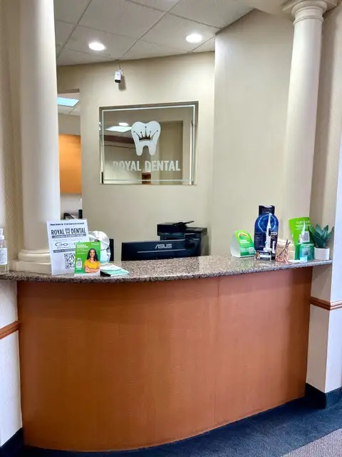 The image shows an interior view of a dental office reception area with a sign for  Royal Dental  on the wall, a counter with items on top, a window, and a person partially visible at the counter.