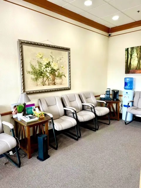 The image shows an interior space with a row of chairs, a framed picture on the wall, and various items on a table.