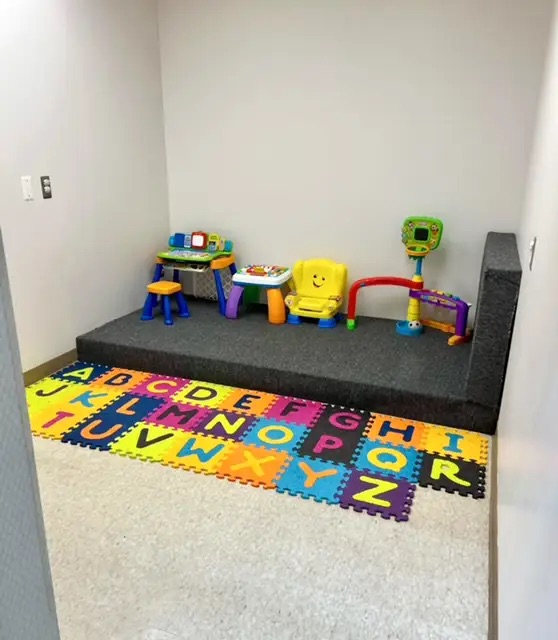 The image shows a room with a play area featuring children s toys, including a carpeted floor with alphabetical letters, numbers, and shapes, and a small table with chairs in front of it. There are also various toys scattered around the area.