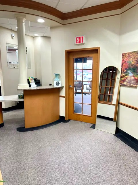 The image shows an interior space with a carpeted floor, a wooden door on the right, a reception desk with a counter and chair, a framed picture hanging above the desk, and a large window allowing natural light into the room.