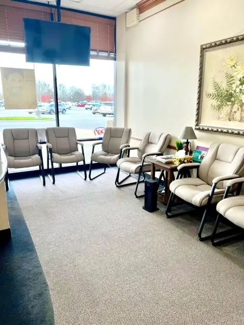 The image depicts an interior space, likely a waiting room, with multiple chairs arranged around a central table, a television mounted on the wall displaying a blank screen, and a reception desk with a nameplate that reads  Jane Doe.