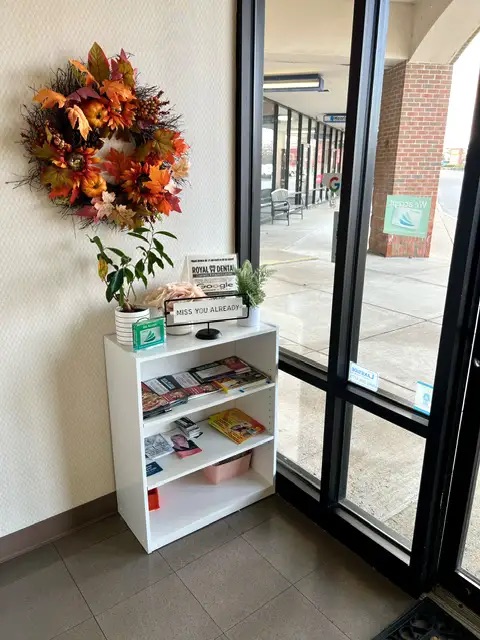 The image shows a cozy corner with a small white table holding a vase with autumn leaves, a framed sign, a stack of books, and a potted plant, all set against a backdrop of a window with a view of a parking lot outside.