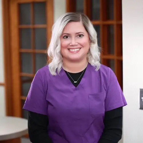 A woman wearing a purple scrub top stands in front of a mirror with a smile on her face.