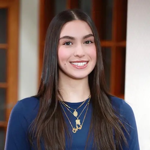 The image shows a young woman with long dark hair smiling at the camera. She is wearing a blue top and has gold jewelry on her necklace.