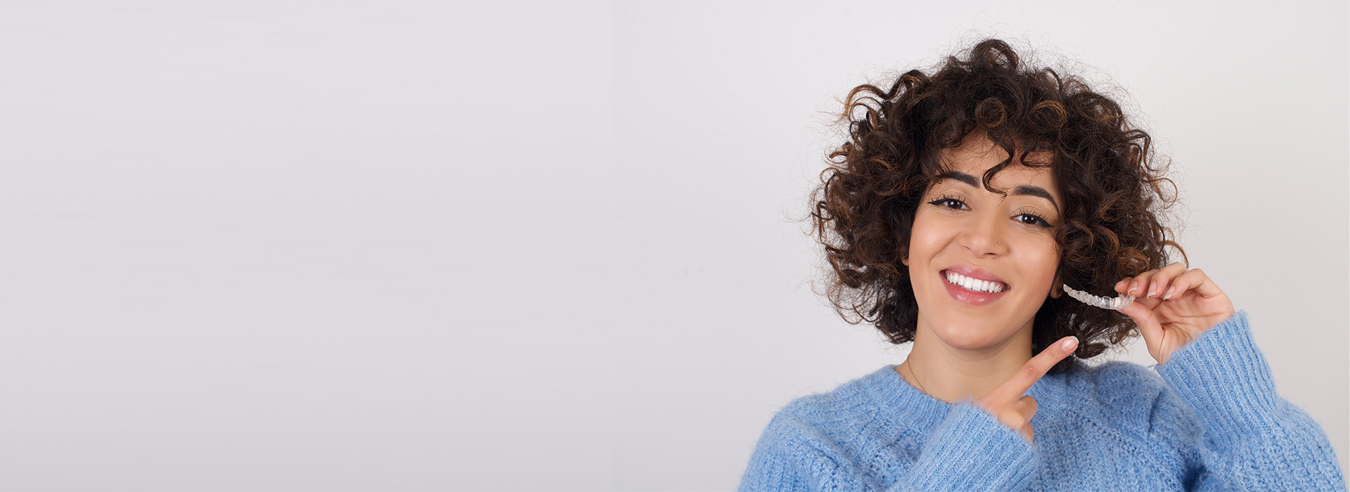 The image shows a woman with curly hair smiling at the camera while holding a toothbrush, set against a plain background.