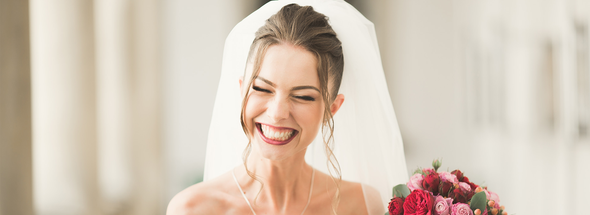 A bride wearing a white veil and holding a bouquet of flowers, smiling at the camera.