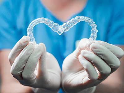 The image shows a person s hands holding a heart-shaped object with a clear, translucent material that appears to be a dental appliance, such as a retainer, against a background that suggests a clean environment, possibly a dental office.