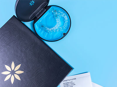 An open cosmetic product box with a blue lid and a clear jar containing a greenish-blue substance on a light blue surface, next to a closed box with a black lid that has a white label with a golden flower design.