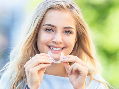The image features a young woman with blonde hair smiling at the camera while wearing clear braces on her teeth.