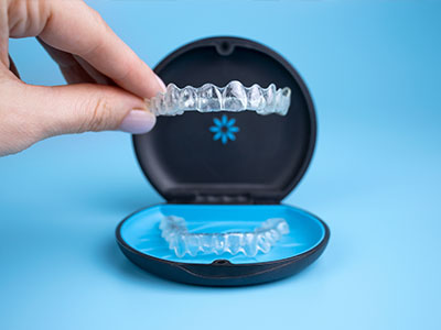 The image shows a person s hand holding a clear plastic tray with a single blue toothbrush inside, placed next to an open black case containing a transparent dental retainer with visible ridges, all set against a light blue background.