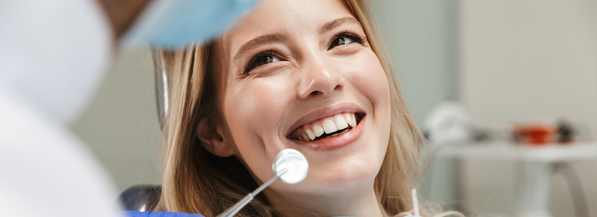 A woman with a bright smile sits in a dental chair, looking up at the camera, while a dentist examines her teeth.