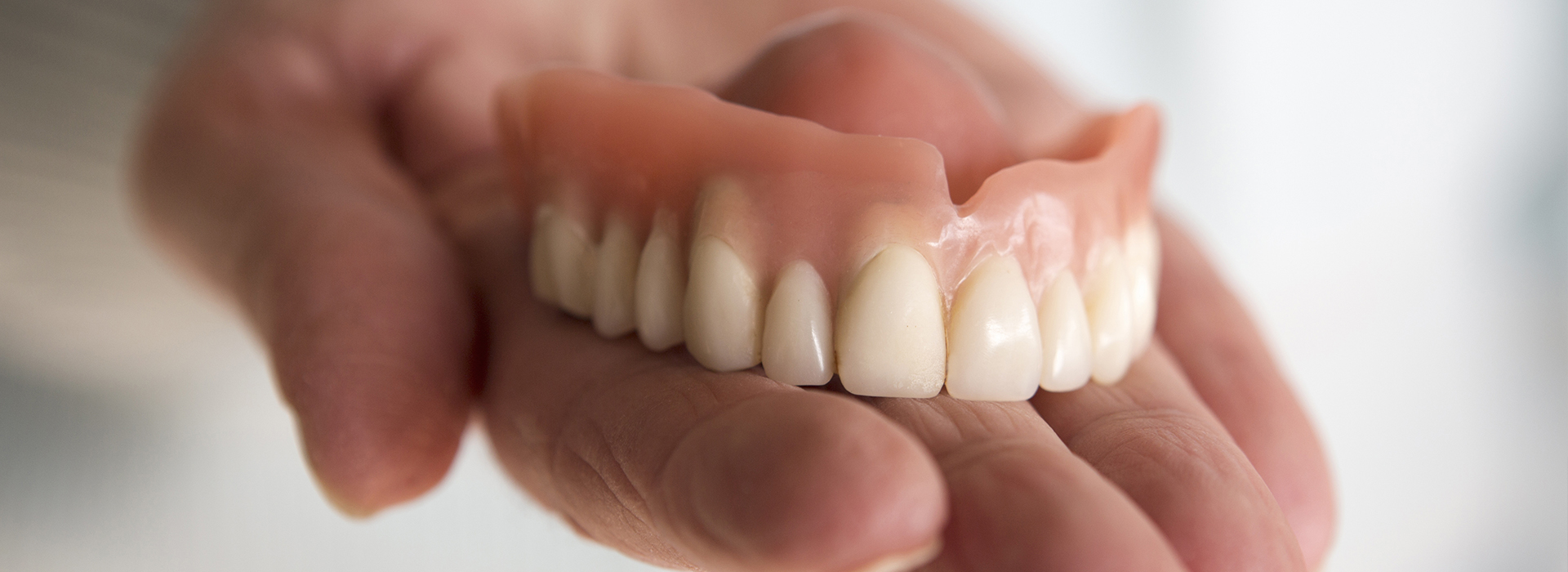 The image shows a person holding a set of dentures against a light background, with a focus on dental hygiene.