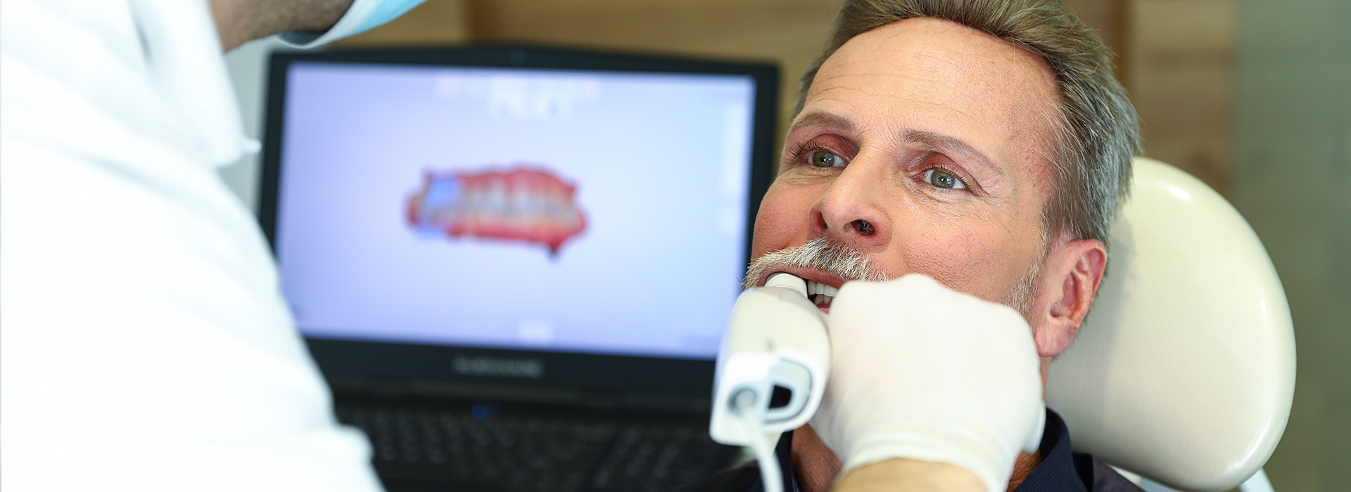 The image depicts a man receiving dental treatment, with a dentist performing a procedure on him while he is seated in a chair with his mouth open, and a computer screen displaying a digital rendering of a tooth in front of them.
