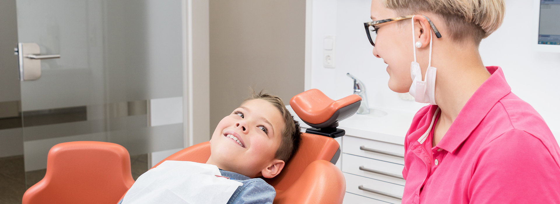 The image shows a dental office setting with a child seated in a dental chair receiving care, a woman standing next to them wearing a face mask and holding an instrument, and another person sitting at a desk observing the situation.