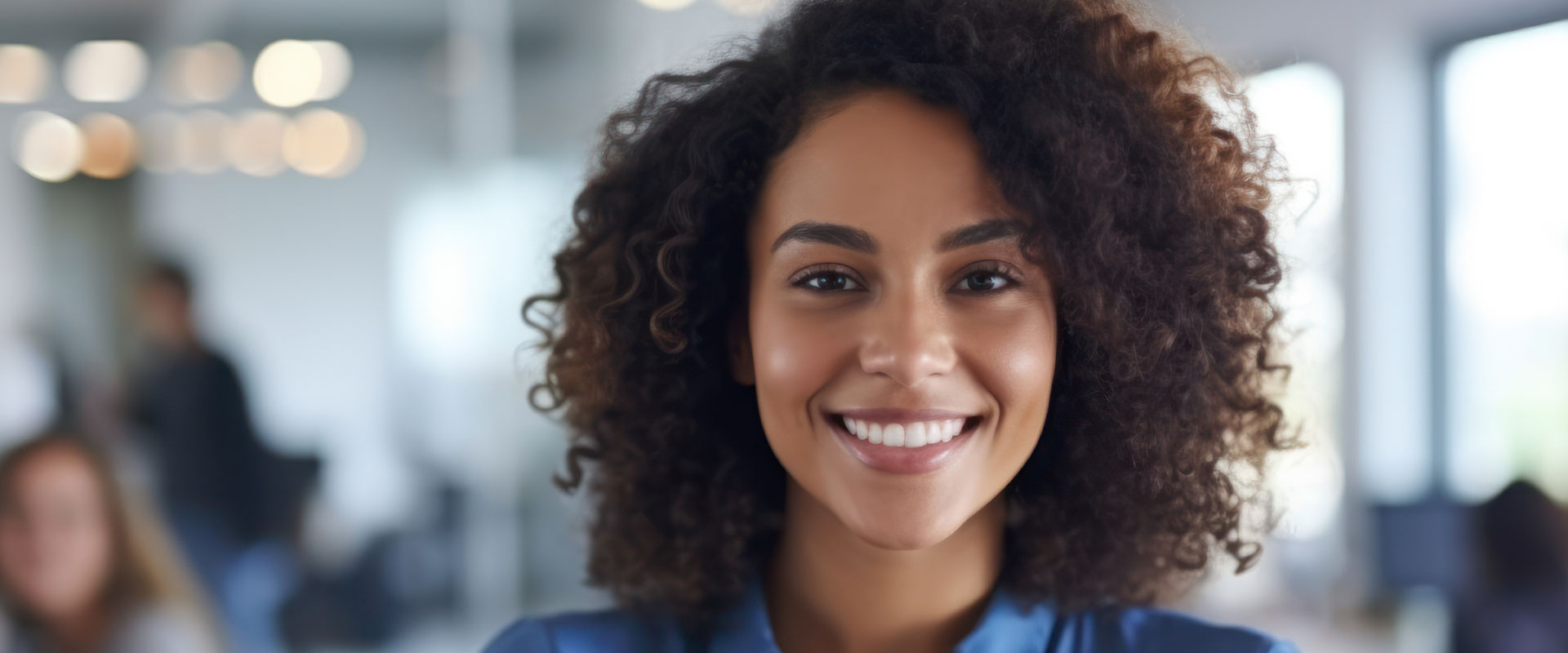 The image shows a smiling woman with curly hair wearing a blue top, standing in an office environment with other people present in the background.