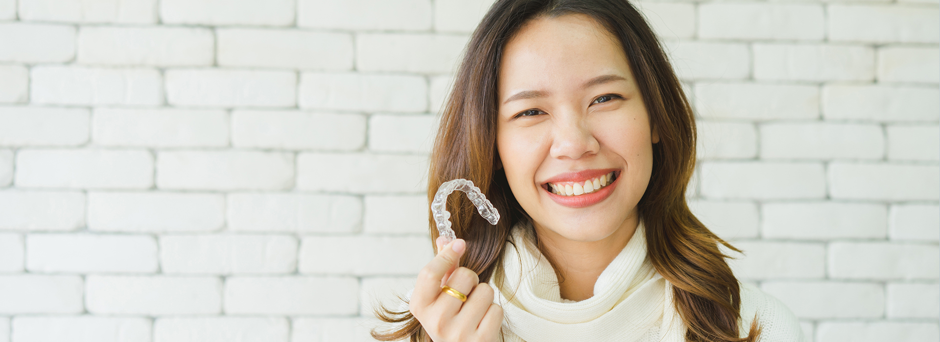 A woman with a smile holds up a toothbrush against her teeth, set against a brick wall background.