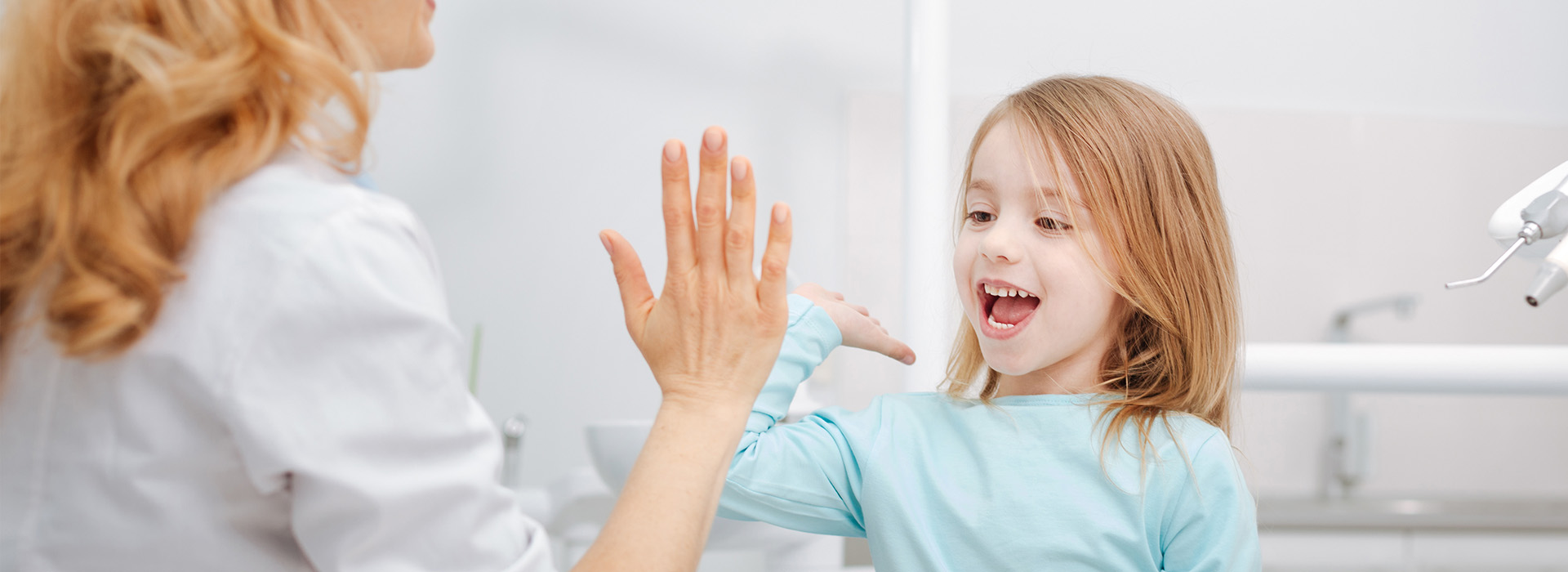 The image depicts a young girl with blonde hair clapping her hands while standing next to an adult woman who appears to be smiling at her. They are both indoors, likely in a bathroom, as suggested by the presence of a sink and a mirror in the background.