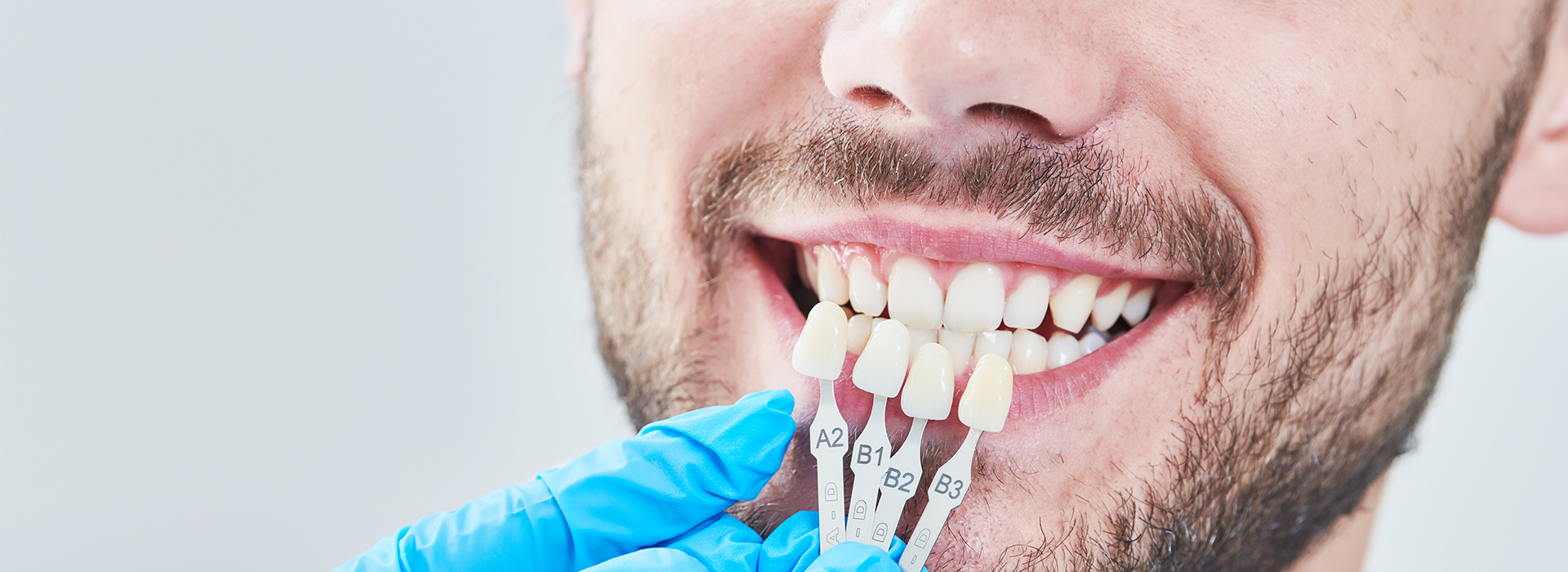 The image shows a person with a wide smile, displaying straight white teeth, holding a toothbrush against their teeth while wearing a blue surgical mask and gloves, with dental tools visible in the background.