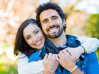 The image features a man and a woman embracing each other with smiles on their faces, set against an outdoor background during daylight.