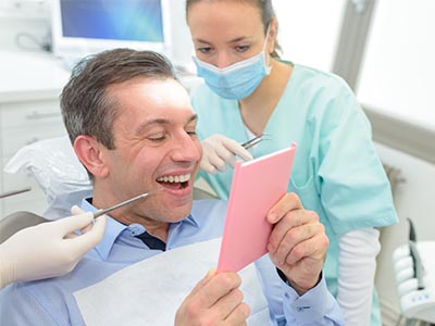 A person is sitting in a dental chair, holding a pink card and smiling at it, while a dental professional looks on with a mask covering their face.