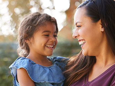A woman and child smiling at the camera, with the adult holding the young one, set against an outdoor backdrop.