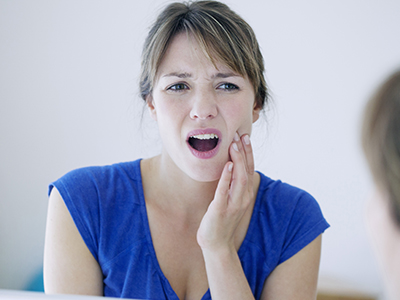 A woman with her mouth open, expressing surprise or shock, looking at her reflection in a mirror while holding her hand to her face.