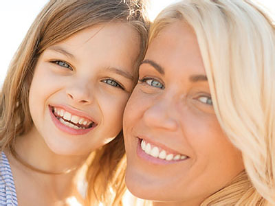 The image shows a woman and a young girl smiling at the camera, with the woman s arm around the child. They appear to be enjoying a sunny day outdoors.