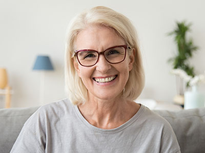 The image depicts a woman with short blond hair, wearing glasses, smiling at the camera. She has light skin, wears a gray top, and is seated indoors with a neutral-toned background featuring a lamp and some decorative items.