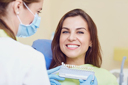 The image shows a dental professional holding a dental implant device while smiling at the camera, with another person sitting next to them wearing protective eyewear and a surgical mask, suggesting a dental care setting.