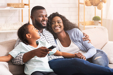 A family of four sitting on a couch, with a man holding a child and smiling at the camera while a woman embraces him and also smiles.