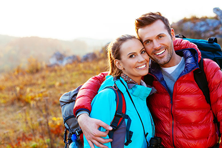 A man and woman are smiling and embracing each other outdoors, with the man wearing a backpack and both dressed for travel, standing on a field of grass at sunset with trees and hills in the background.