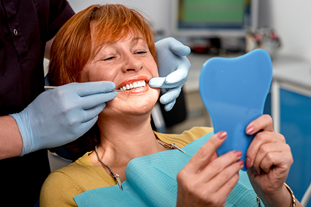 A woman sitting in a dental chair with a smile on her face, holding up a blue toothbrush-shaped object in front of her while a dental professional works on her teeth.