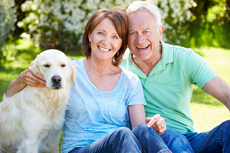 A man and woman are sitting outdoors with a dog between them  they all appear relaxed and happy, with the man smiling at the camera.