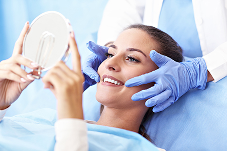 A woman receiving a facial treatment with a large magnifying glass held over her face by a professional in a medical setting, smiling at the camera.