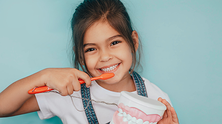 A young girl brushing her teeth with a toothbrush, smiling at the camera, holding a red toothbrush, standing in front of a blue wall.