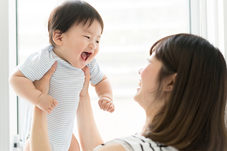 The image depicts a woman holding a baby who appears to be laughing or crying with joy, while another adult looks on with a smile. They are indoors with natural light coming through a window.