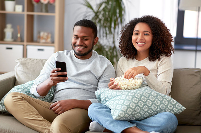 Two people sitting on a couch, sharing a moment with snacks while watching something together on a phone screen.