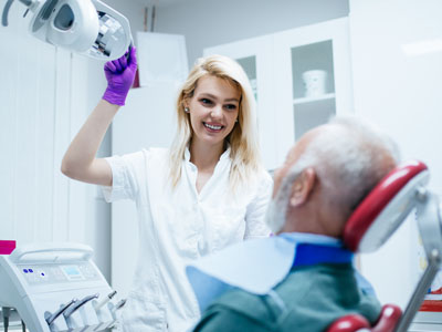 The image shows a woman standing next to an older man who appears to be undergoing dental treatment, with the woman holding a mirror up to the man s face, possibly assisting or examining him during the procedure.