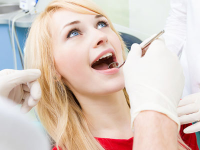 The image shows a dental office setting where a woman with blonde hair is seated in a dental chair, smiling broadly while undergoing a dental procedure  a dentist is visible, wearing gloves and working on her teeth, with dental tools and equipment around them.