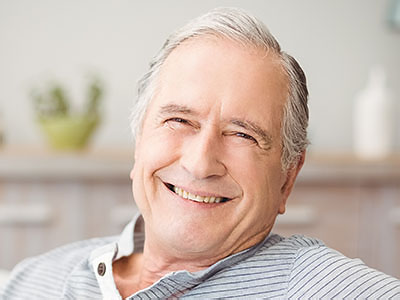 An elderly man with gray hair, smiling broadly while seated in a chair indoors.