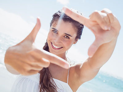 A young woman with long hair and a bright smile takes a selfie with her hands framing the camera lens while standing on a beach, with the ocean visible behind her.