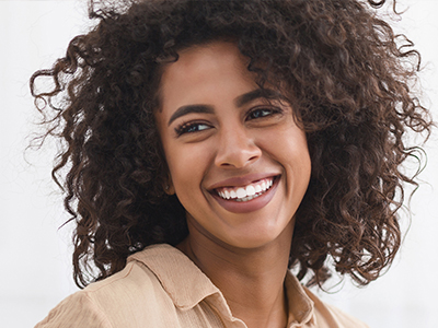 A smiling woman with curly hair, wearing a beige top, against a blurred background.